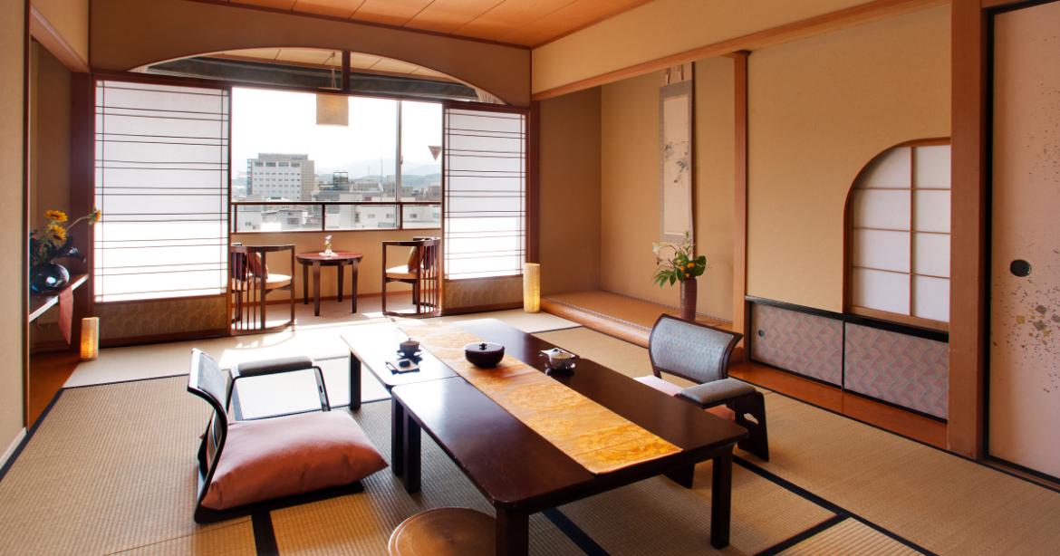 Camera in stile tradizionale giapponese (ryokan) con pavimentazione in tatami, tavolo basso da tè, sedie da terra e porte scorrevoli shoji aperte sulla vista della città