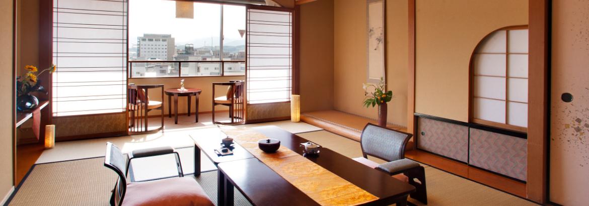 Camera in stile tradizionale giapponese (ryokan) con pavimentazione in tatami, tavolo basso da tè, sedie da terra e porte scorrevoli shoji aperte sulla vista della città